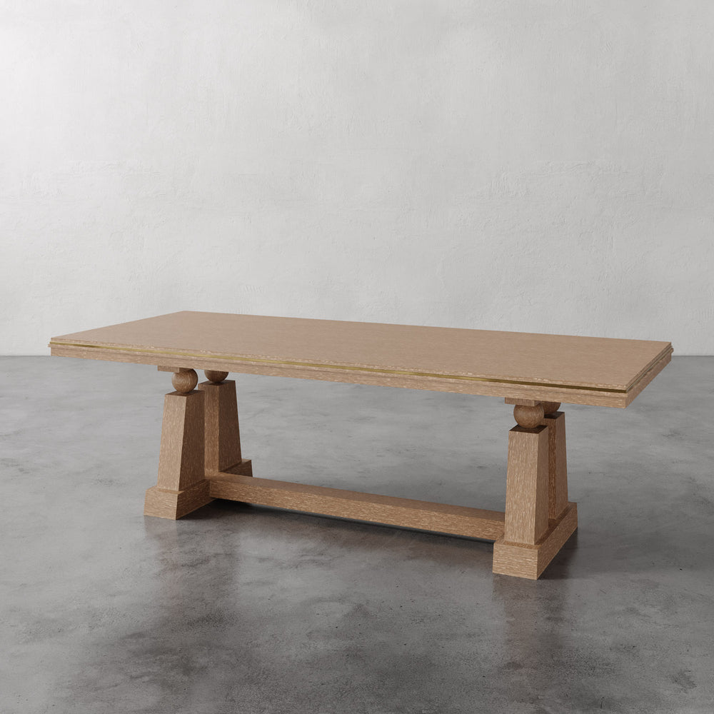 Lemieux et Cie Corvaux Dining Table in warm oak, dining table with sculptural trestle base, shown on polished concrete floor against textured plaster walls.