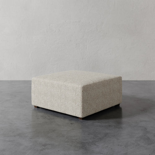 Lemieux et Cie Fabien 36" Square Ottoman in textured ivory bouclé, low-profile upholstered ottoman on stone floor against textured plaster walls.