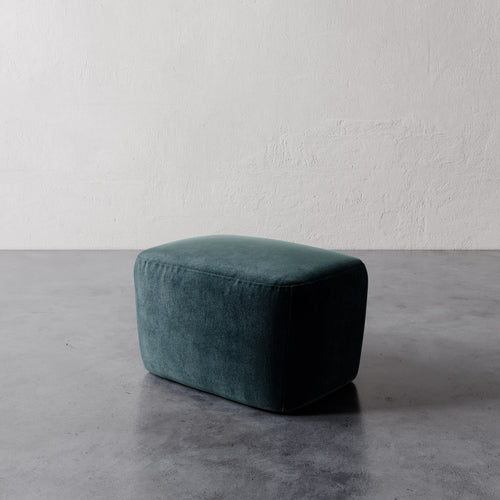 Lemieux et Cie Aurélie Ottoman in deep teal, rounded low-profile ottoman with soft upholstery, shown on polished concrete floor against textured plaster wall.