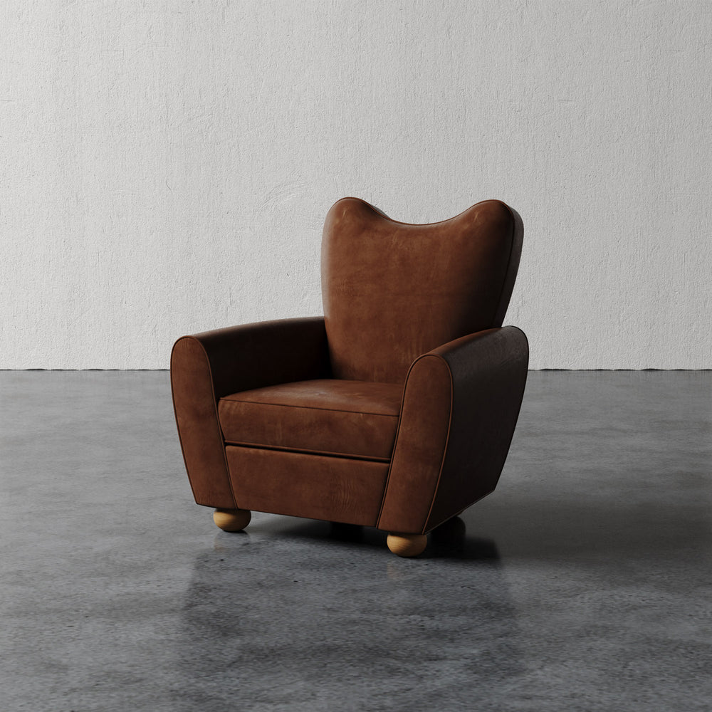 Lemieux et Cie Danielle Chair in tobacco brown leather with sculptural curved backrest and turned wood feet, shown on concrete floor against white plaster wall.