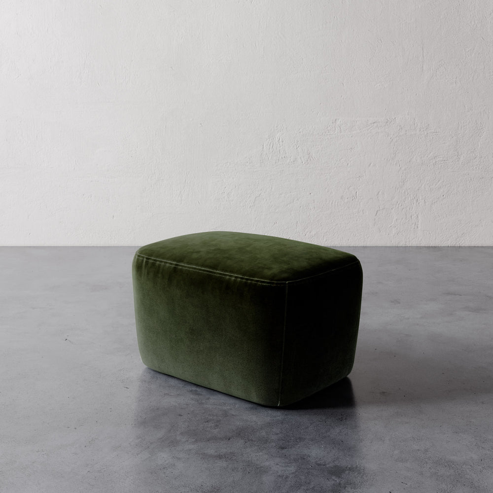 Lemieux et Cie Aurélie Ottoman in deep green, rounded low-profile ottoman on polished concrete floor with textured white plaster wall.
