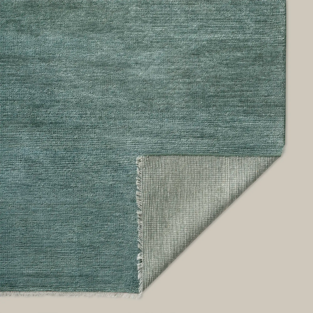 Lemieux et Cie Rochefort Handmade Wool Rug in soft teal, hand-knotted rug with subtle fringe detail, corner folded to reveal textured backing on neutral floor.