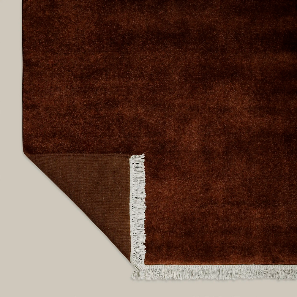 Lemieux et Cie Plume Handmade Wool Rug in oxblood, folded corner detail showing woven backing and white fringe on neutral backdrop.