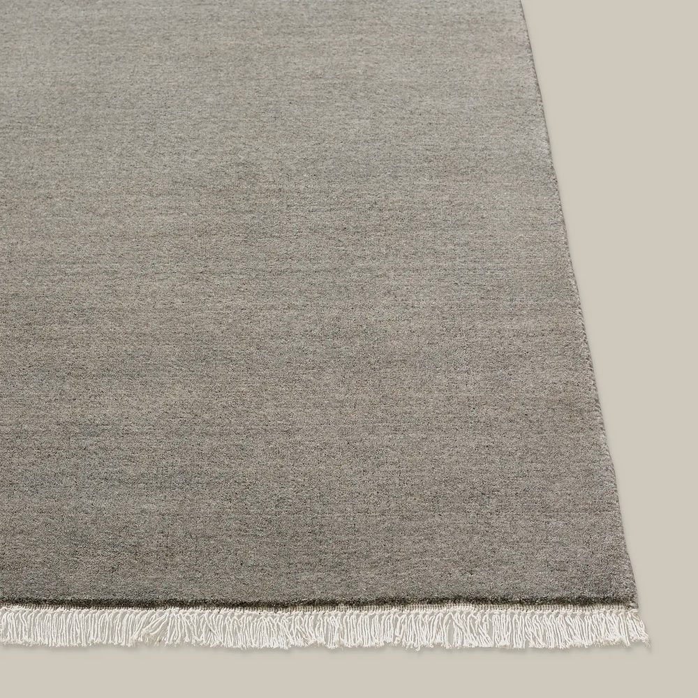 Lemieux et Cie Plume Handmade Wool Rug in grey, textured solid rug with hand-knotted fringe, shown on neutral surface in soft light.