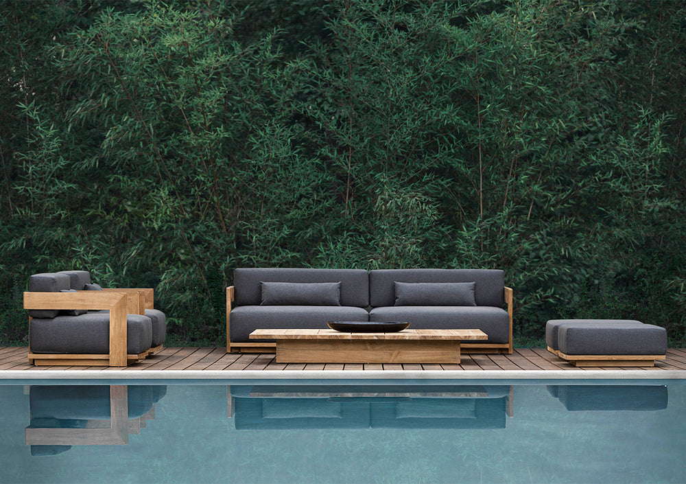Lemieux et Cie S2 Teak Ottoman in charcoal upholstery with solid teak base, outdoor seating styled on wood deck beside pool and greenery.