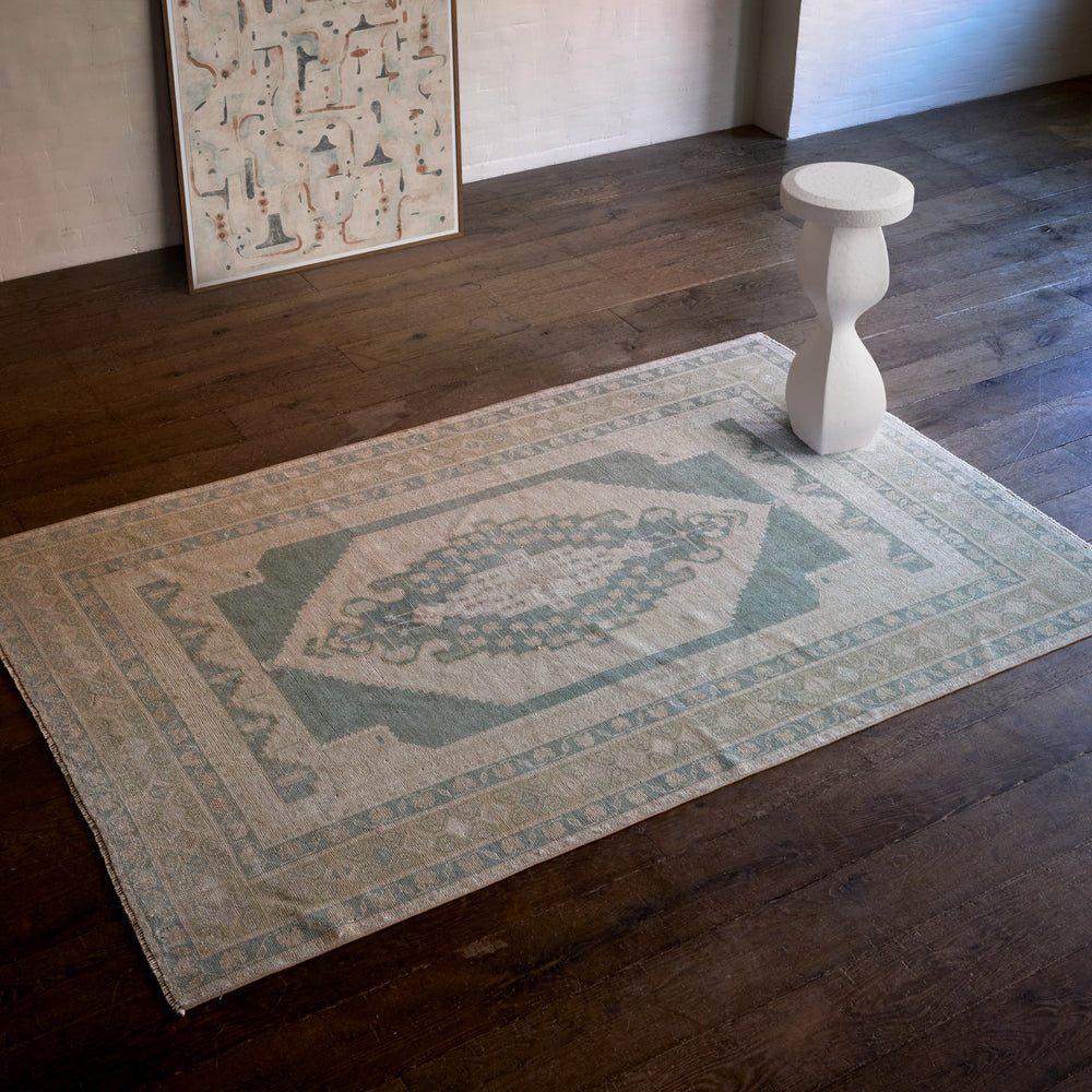 Lemieux et Cie Vintage 1960's Anatolian rug in faded sage and warm neutrals, handwoven vintage carpet on dark wood floors beside sculptural plaster side table.