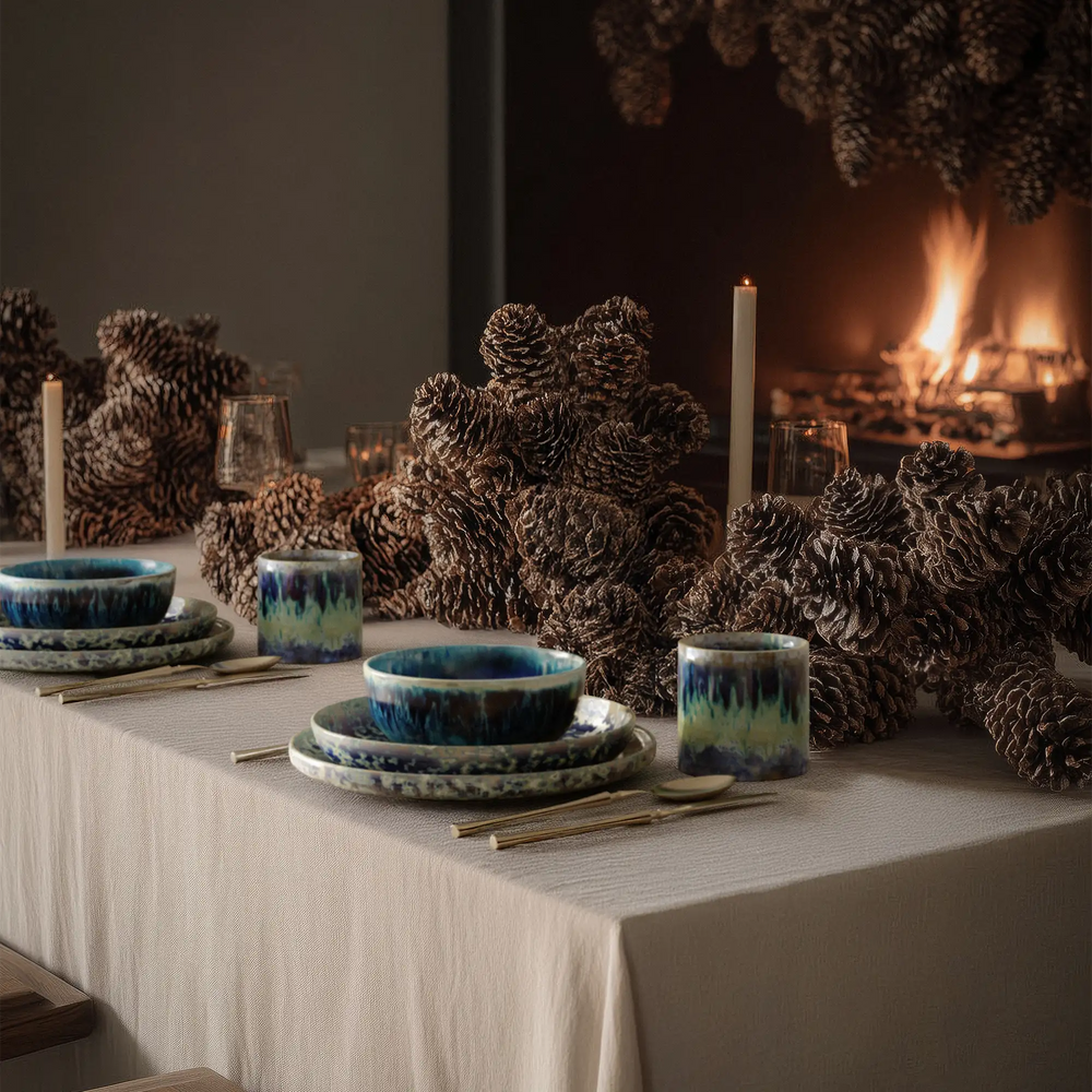 Lemieux et Cie Hanami Stoneware Bowl Set in Indigo, ceramic bowls with reactive glaze, styled on linen-covered table with pinecones, candles, and fireplace glow.