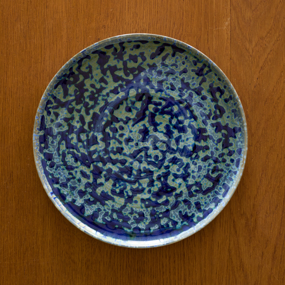 Lemieux et Cie Hanami Stoneware Dinner Plate in indigo with reactive glaze, shown overhead on warm wood surface with organic tonal variation.