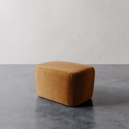 Lemieux et Cie Aurélie Ottoman in warm caramel, rounded upholstered ottoman on polished concrete floor with textured white plaster walls