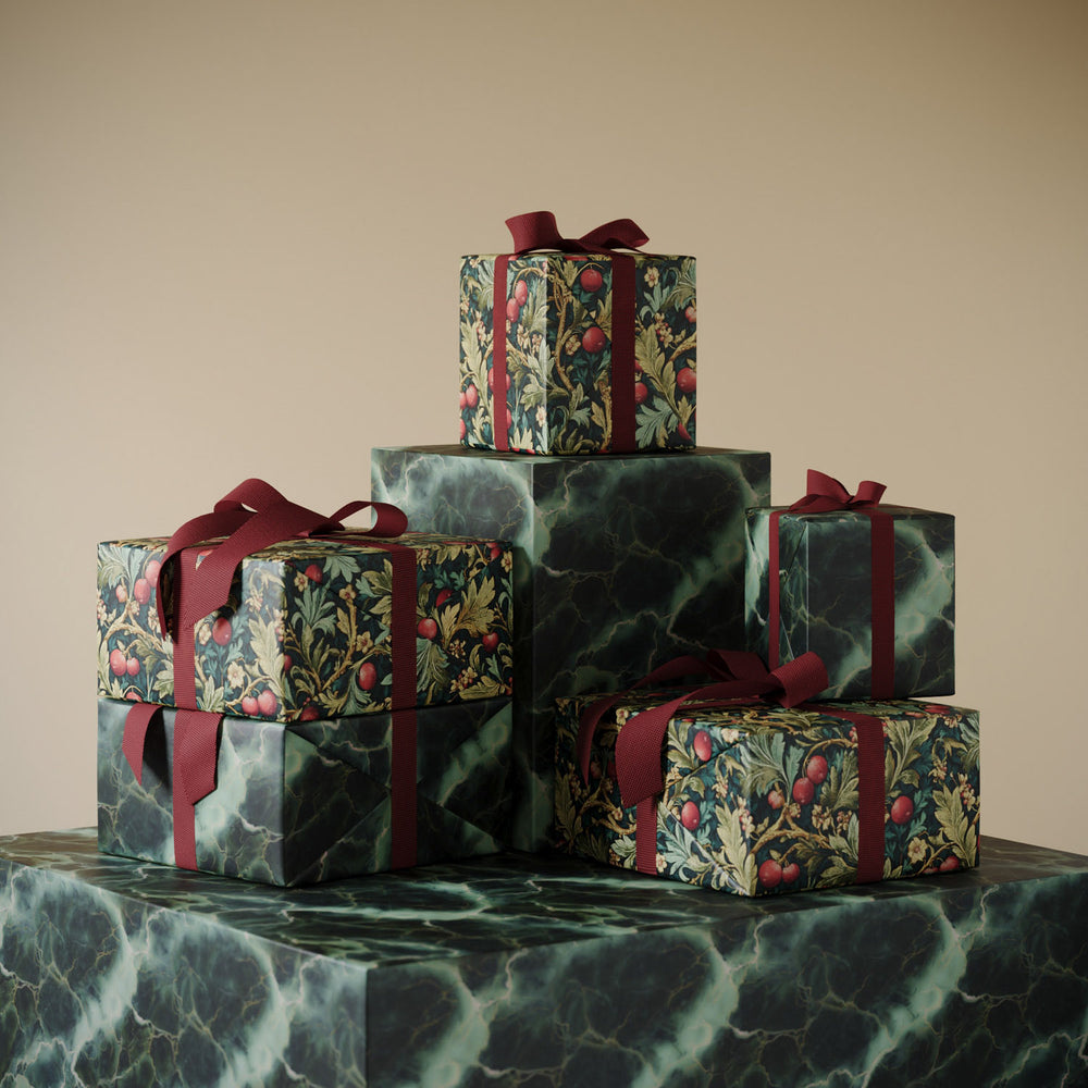 Lemieux et Cie Holly Berries Gift Wrapping Paper with botanical motif and burgundy ribbon, stacked boxes on green veined marble table against neutral backdrop.