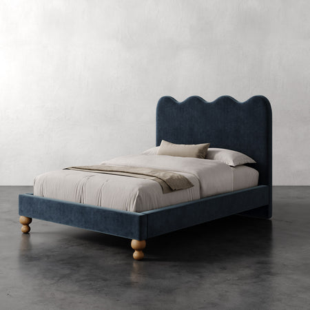 Lemieux et Cie Celine Bed with scalloped headboard in deep blue, upholstered bed with turned legs on concrete floor against textured plaster walls.