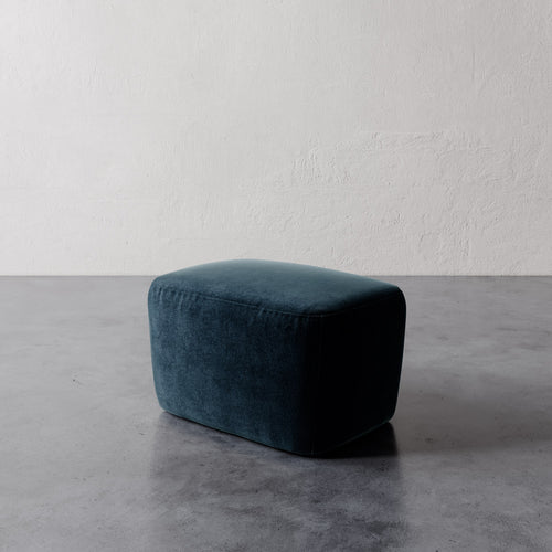 Lemieux et Cie Aurélie Ottoman in deep teal, rounded upholstered pouf shown on concrete floor against textured plaster walls.