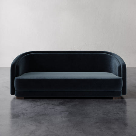 Lemieux et Cie Valence Sofa in deep indigo, rounded low-profile sofa on dark legs, shown on concrete flooring against neutral plaster wall.