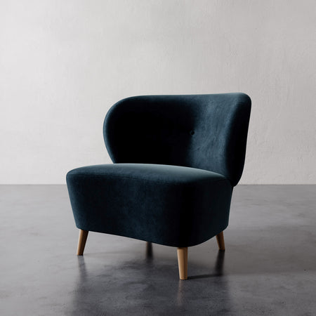 Lemieux et Cie Tourenne Chair in deep teal velvet upholstery with tapered legs, organic rounded silhouette on concrete floor against textured plaster wall.