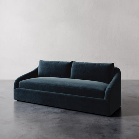 Lemieux et Cie Rousseau Sofa in deep teal upholstery, curved-arm sofa with bench seat cushion, shown on concrete floors against textured plaster walls.