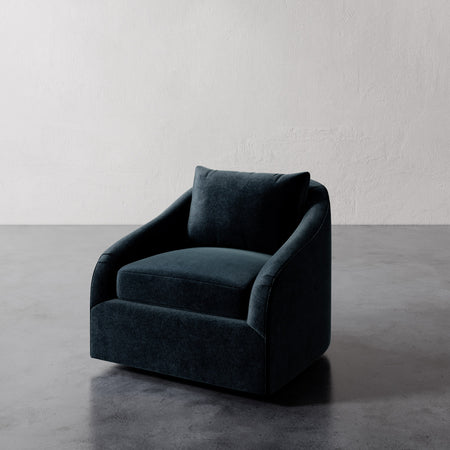 Lemieux et Cie Rousseau Swivel Chair in deep teal upholstery, curved arm lounge chair on concrete floor against white textured walls.