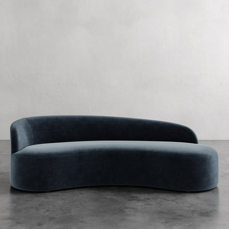 Lemieux et Cie Colette Left Arm Sofa in deep blue-green upholstery, curved asymmetric sofa on concrete floor against neutral plaster wall.