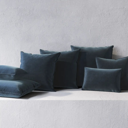 Lemieux et Cie throw pillows in deep teal with welted edges, soft upholstered cushions in square and lumbar forms, arranged on textured plaster shelf.