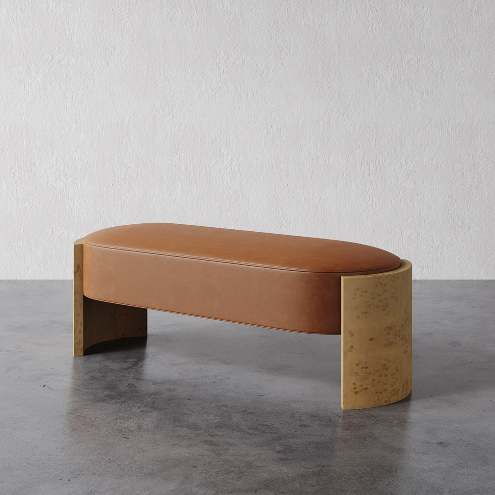 Lemieux et Cie Benoit Bench with oval upholstered top and curved slab supports, warm cognac tone, shown on concrete floor against textured white wall.