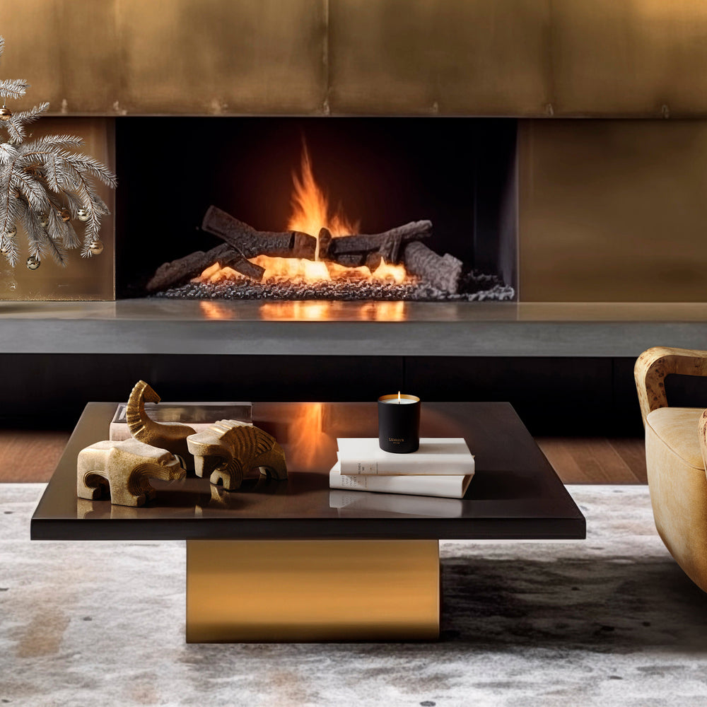 Lemieux et Cie coffee table with brass base and dark top, styled with sculptural animal figures, candle and books, before a stone fireplace in a textured sitting room.