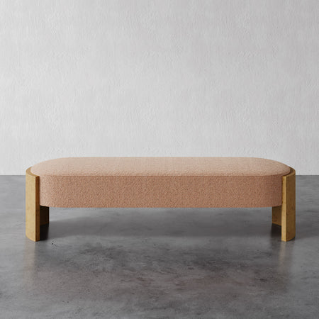 Lemieux et Cie Benoit Bench in warm blush upholstery with blonde curved legs, low-profile bench shown on concrete floor against textured white plaster wall.