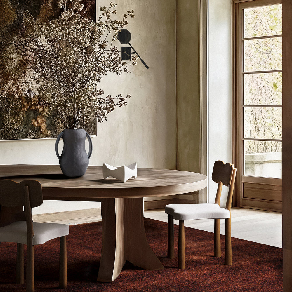 Lemieux et Cie Plume Handmade Wool Rug in oxblood, shown beneath a round dining table with upholstered chairs in a neutral dining room with stone floors.
