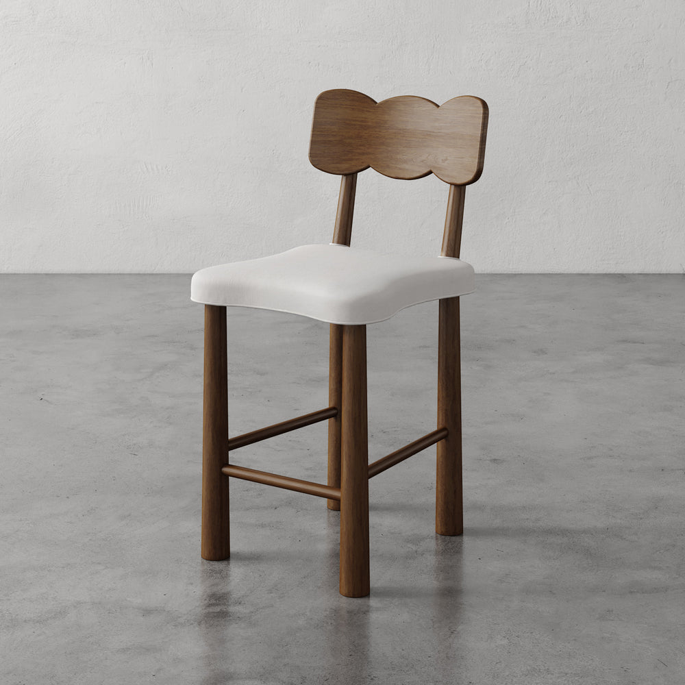 Lemieux et Cie Madeleine Counter Stool, counter-height stool with sculptural backrest and cream upholstered seat, shown on polished concrete floor against textured plaster walls.
