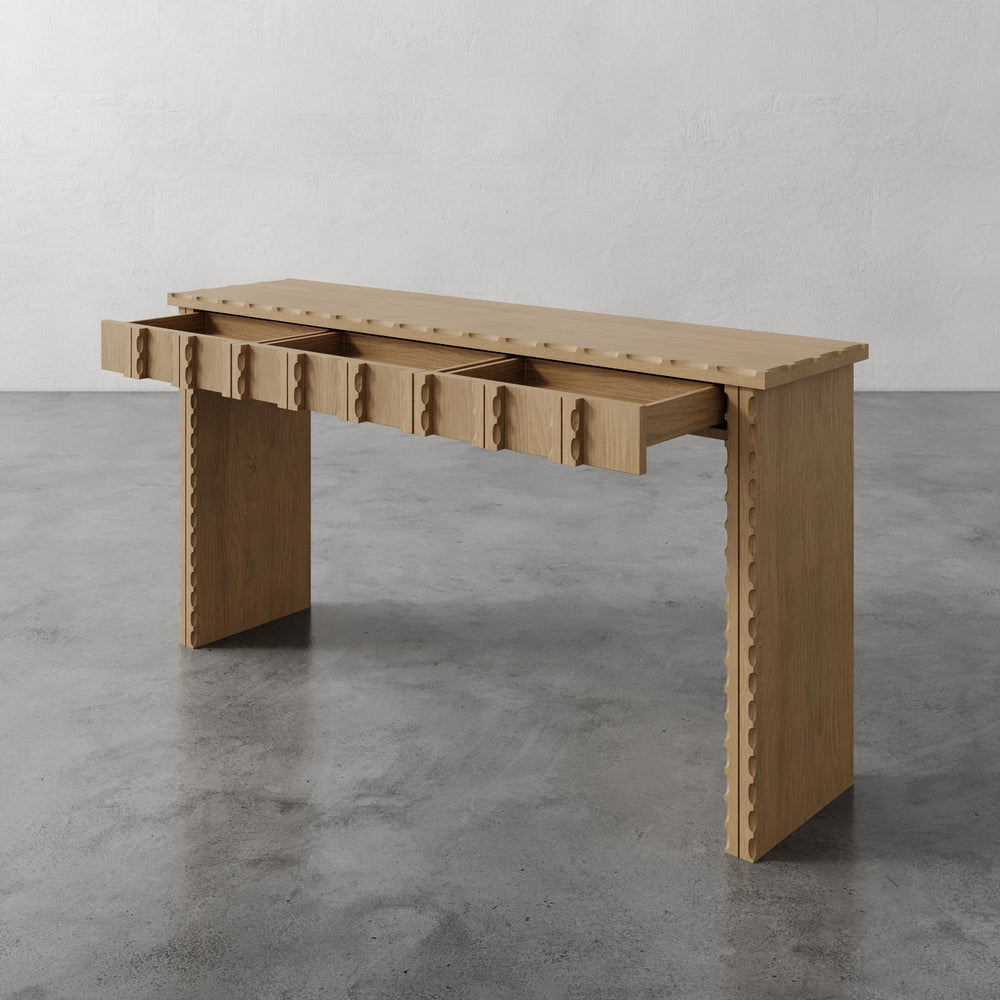 Lemieux et Cie Marchand Console Table in warm oak with scalloped drawers, console table shown on polished concrete floor against white textured walls.