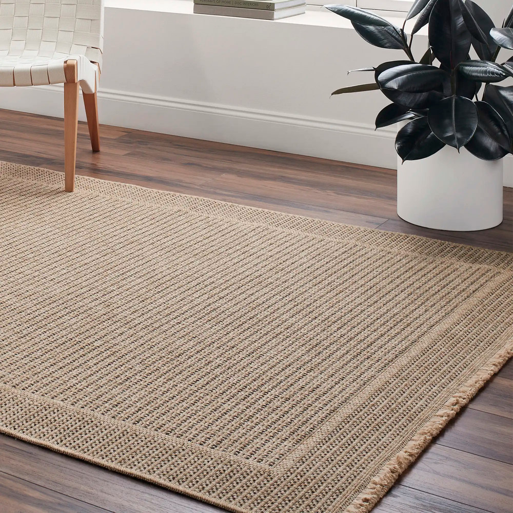 Lemieux et Cie Moreau Outdoor Rug in natural tones, textured flatweave rug with braided border, shown on wood flooring near bench and planter.