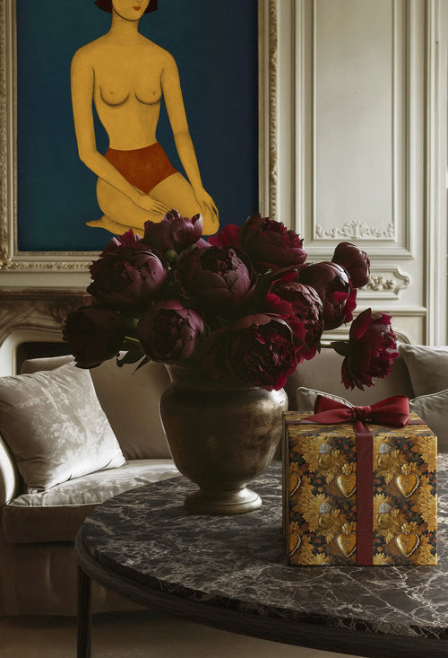 Lemieux et Cie Victorian Locket fancy gift wrapping applied on a box resting on the Ravelle coffee table in a living room with red flowers and the Les femmes artwork in the background