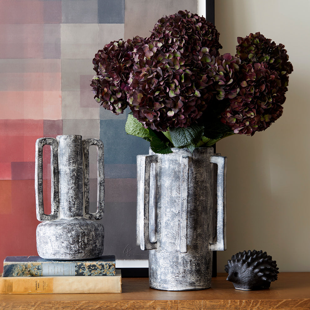 Lemieux et Cie Normandie Vase in light grey washed finish, geometric sculptural vase with square handles, styled with dried hydrangeas on wood surface with colorful abstract artwork and books.