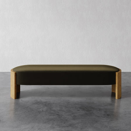Lemieux et Cie Benoit Bench, upholstered bench with olive-toned cushion and curved corner legs, shown on concrete floor against white textured wall.