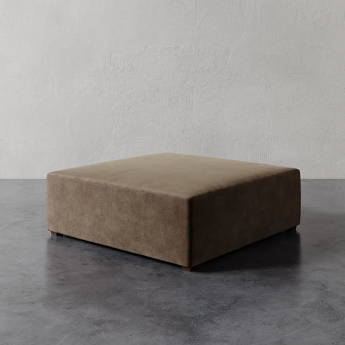 Lemieux et Cie Fabien 48" Square Ottoman in warm taupe, low-profile upholstered ottoman on stone floor against textured plaster wall.