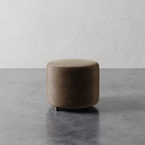 Lemieux et Cie Fabien 22" Round Ottoman, cylindrical upholstered ottoman in warm taupe, shown on gray concrete floor against textured plaster wall.