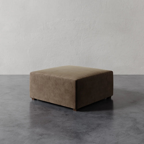 Lemieux et Cie Fabien 36" Square Ottoman in taupe upholstery, low-profile ottoman on dark stone flooring against textured plaster walls.