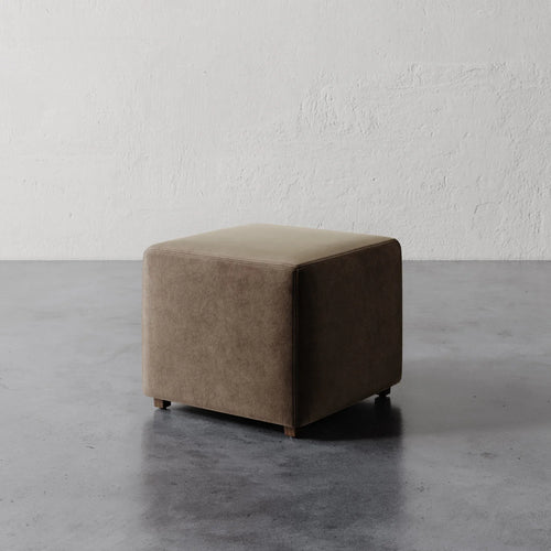 Lemieux et Cie Fabien 22" Square Ottoman in warm taupe upholstery, cube-form ottoman on concrete floor against textured plaster wall.