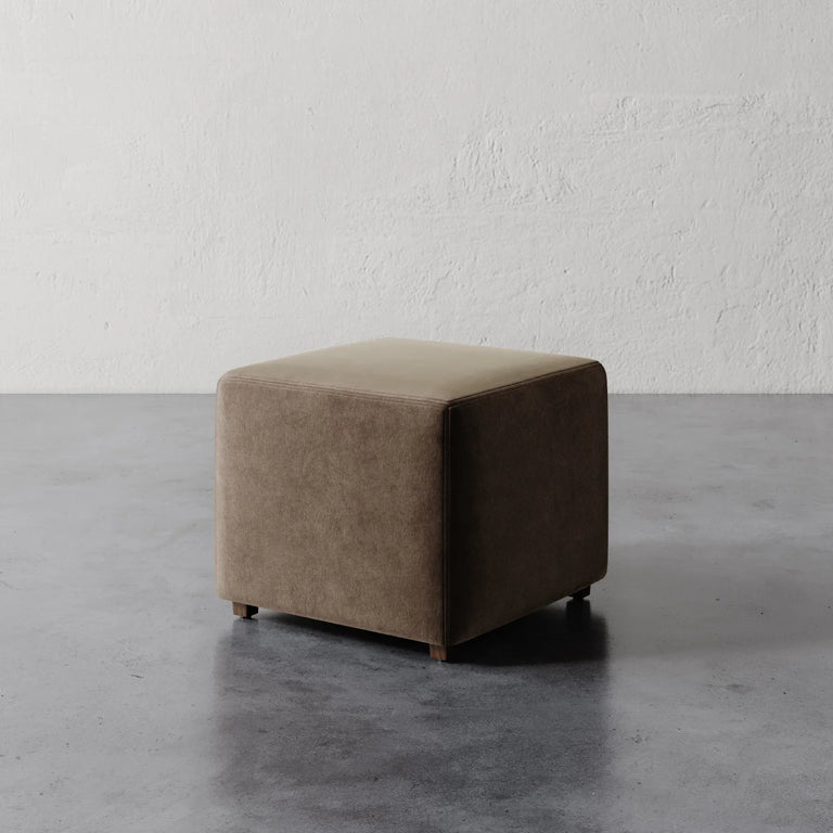 Lemieux et Cie Fabien 22" Square Ottoman in warm taupe upholstery, cube-form ottoman on concrete floor against textured plaster wall.