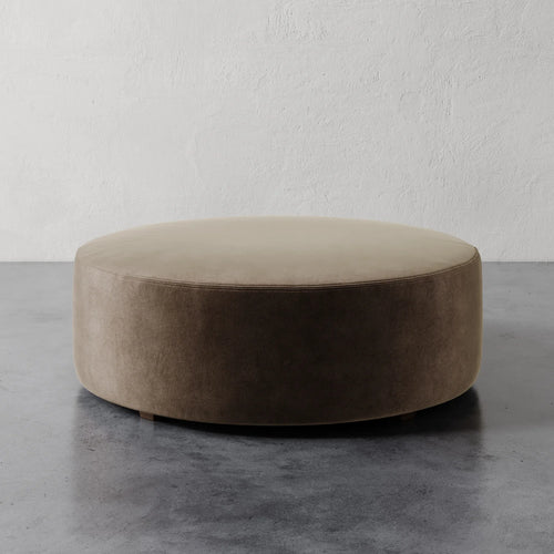 Lemieux et Cie Fabien 48" Round Ottoman, cylindrical upholstered ottoman in warm taupe, shown on polished concrete floor against textured white plaster wall.