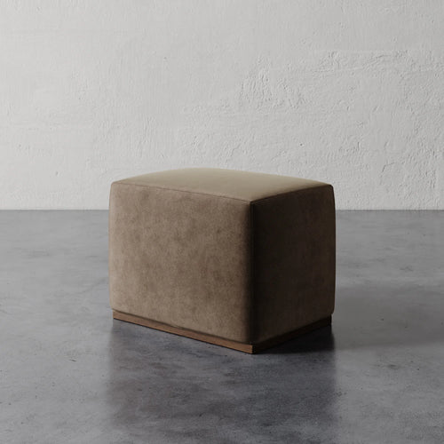 Lemieux et Cie Sophie Rectangular Ottoman in taupe upholstery, low-profile cushioned ottoman on dark base, shown on polished concrete floor against textured white plaster wall.