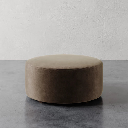 Lemieux et Cie Fabien 36" Round Ottoman in warm taupe upholstery, low-profile round ottoman on polished concrete floor against textured plaster walls.