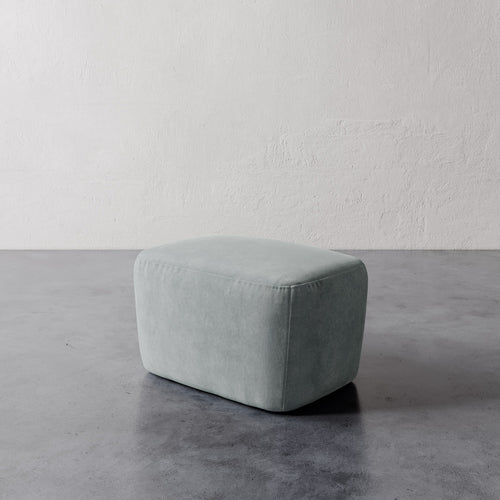 Lemieux et Cie Aurélie Ottoman in soft sage, rounded low-profile pouf shown on concrete floor against textured plaster walls.