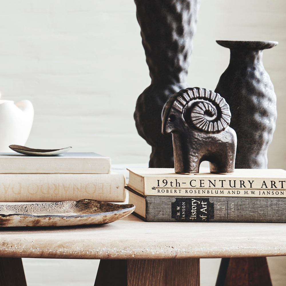 Lemieux et Cie Ram sculptural figurine in textured black glaze, hand-modeled ceramic piece styled on stone table with books and dark vases against neutral wall.