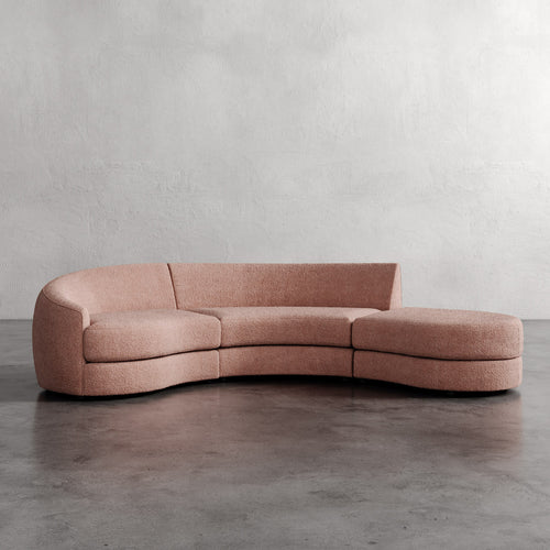 Pink curved sofa on a gray floor with a plain wall background. Rulmann 3-Piece Left Sectional Rosewood Berber