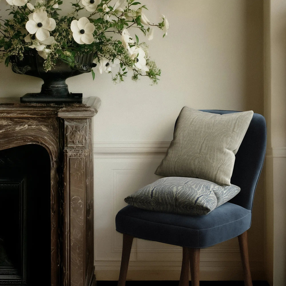 Blue velvet Rozette chair with 2 Linen accent pillows in the cascading willow botanical pattern in neutral and indigo next to a marble mantle with flowers
