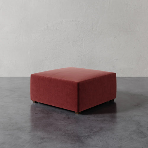Lemieux et Cie Fabien 36" Square Ottoman in terracotta, low-profile upholstered ottoman, shown on polished concrete floor against textured plaster wall.