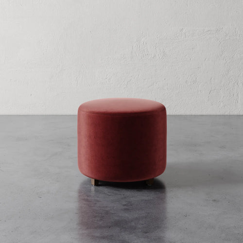 Lemieux et Cie Fabien 22" Round Ottoman in terracotta upholstery, cylindrical accent piece on stone floor against textured plaster wall.
