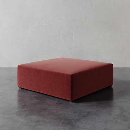 Lemieux et Cie Fabien 48" Square Ottoman in terracotta upholstery, low-profile ottoman on dark concrete floor against textured plaster walls.