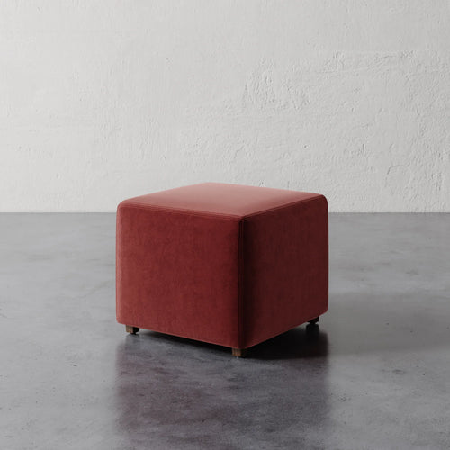 Lemieux et Cie Fabien 22" Square Ottoman in terracotta upholstery, low-profile cube-shaped seating on concrete floor against textured white plaster wall.