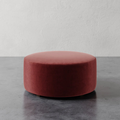 Lemieux et Cie Fabien 36" Round Ottoman in warm terracotta upholstery, low-profile circular ottoman on polished concrete floor against textured white plaster wall.