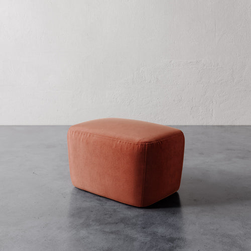 Lemieux et Cie Aurélie Ottoman in warm terracotta, low-profile rounded cube ottoman on polished concrete floor against textured plaster wall.